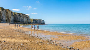 Plage de galets et falaises de Fécamp sous ciel bleu. Des promeneurs profitent du soleil au bord de la mer.