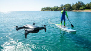 Drone gris HoverAir Aqua en vol au-dessus de l'eau claire, filmant un homme en combinaison de plongée faisant du stand-up paddle près d'une plage tropicale.
